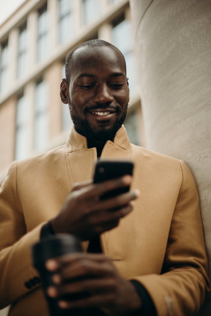 A cheerful man in a stylish coat using his smartphone and holding coffee outside.