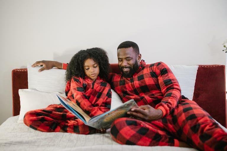 A father and daughter in red plaid pajamas reading a bedtime story together on a bed.