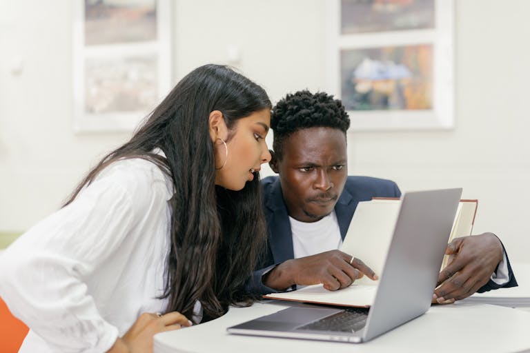 A focused discussion between two adults using a laptop and book in an indoor setting.
