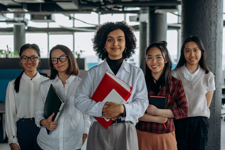 Group of diverse women colleagues smiling in a modern office setting, holding documents.