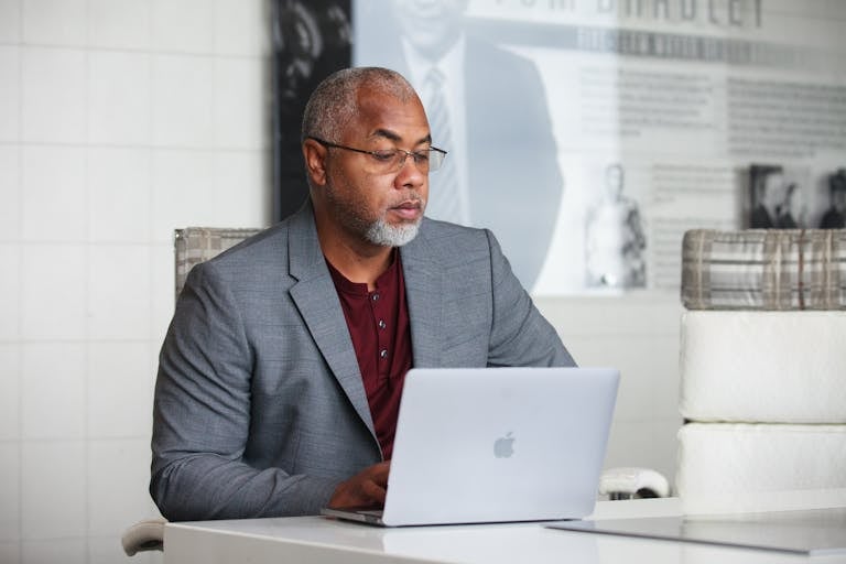 Senior adult man with gray beard working remotely on laptop indoors wearing a blazer.