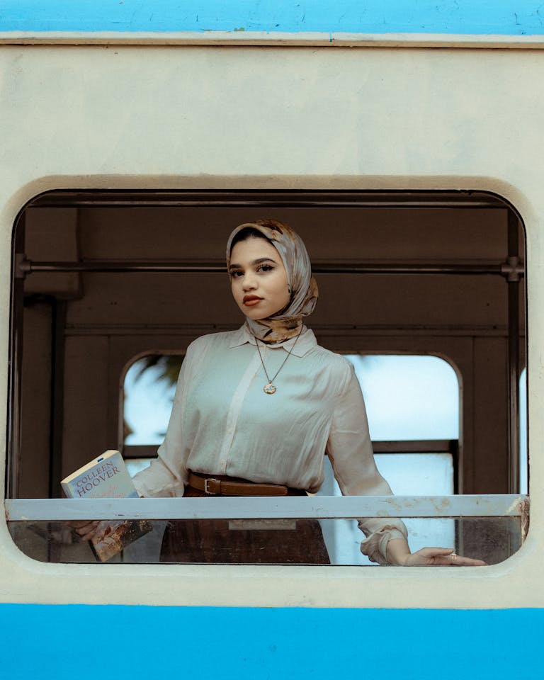 Young woman in hijab holding a book, seen through a train window in Egypt.