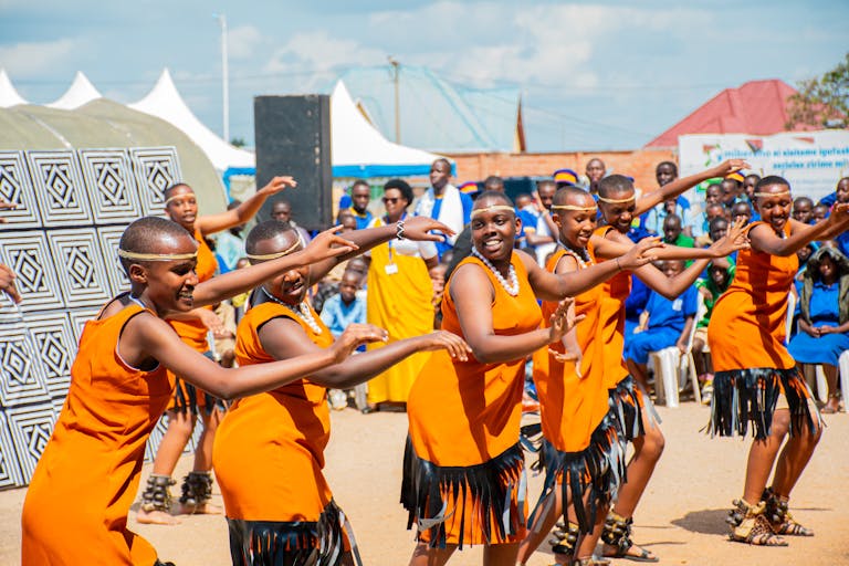 Group of African women performing traditional dance in vibrant orange dresses outdoors.