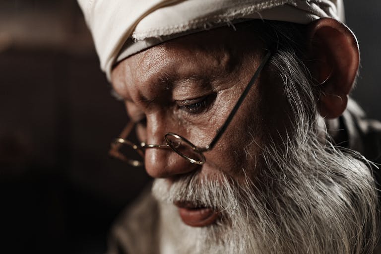 Close-up portrait of an elderly man wearing glasses and a white headscarf, deep in thought.